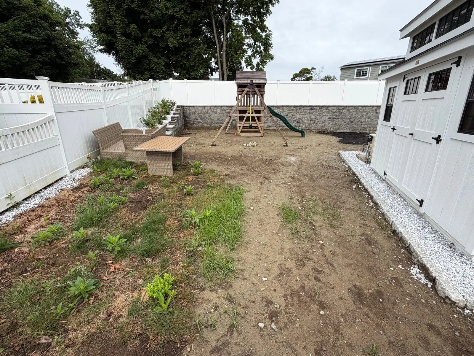 The bare backyard in Milford, CT before sod — graded dirt around a wooden playset, bordered by a white vinyl privacy fence, a stacked-stone wall, and a board-and-batten outbuilding