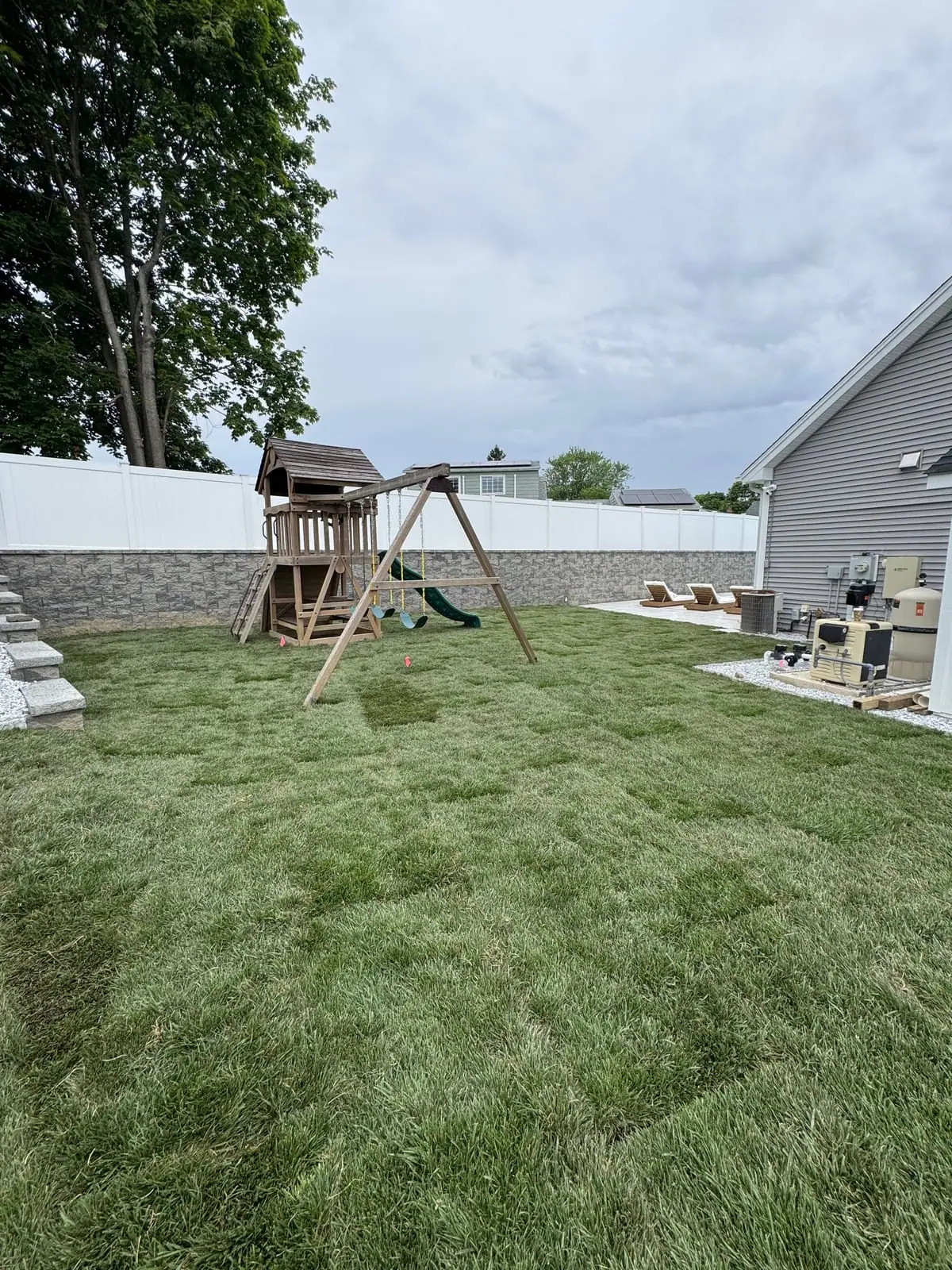 Finished tall fescue backyard lawn around a wooden playset in Milford, CT, bordered by a white privacy fence with the pool patio and lounge chairs along the house