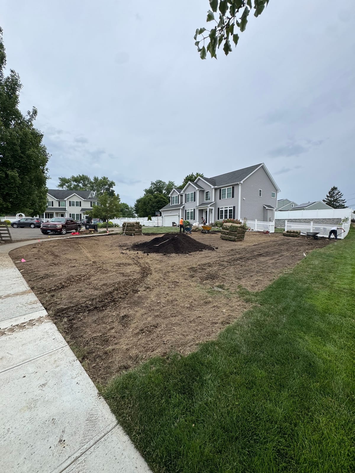 The stripped front yard in Milford, CT on install day — bare graded soil with a pile of dark topsoil, stacks of tall fescue sod pallets, and the crew starting to lay rolls up by the gray colonial