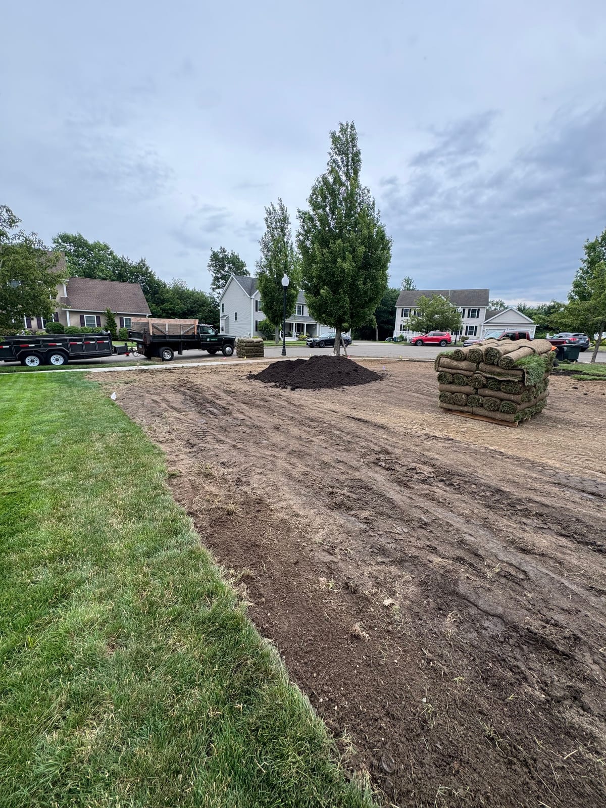 Pallets of fresh tall fescue sod and a pile of dark topsoil staged on the stripped, graded front yard in Milford, CT, with the crew's dump truck and trailer parked on the cul-de-sac behind