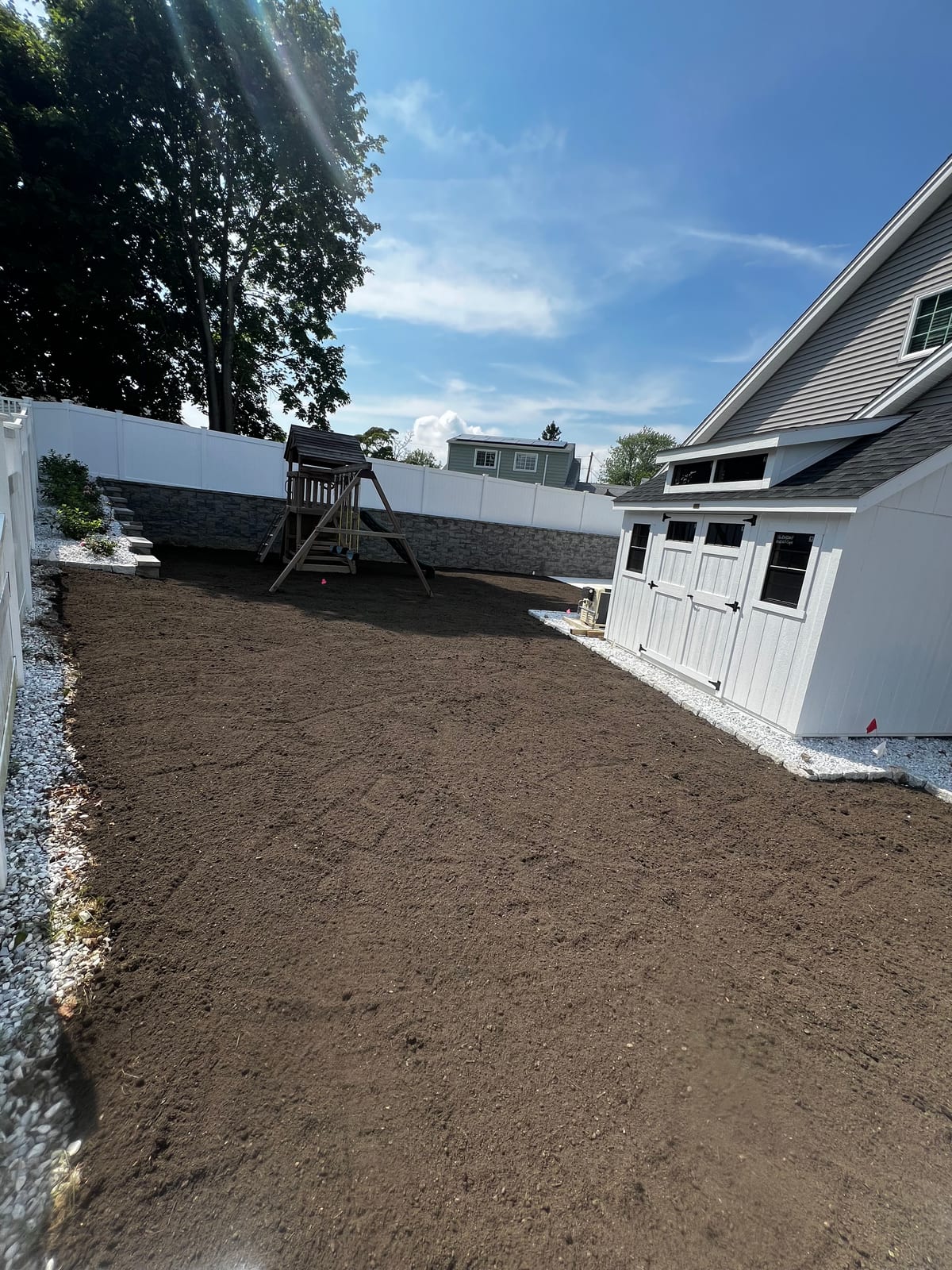 The graded Milford, CT backyard ready for sod — a wide, level bed of raked dirt running past a wooden playset and a stacked-stone wall on one side and a white board-and-batten shed on the other