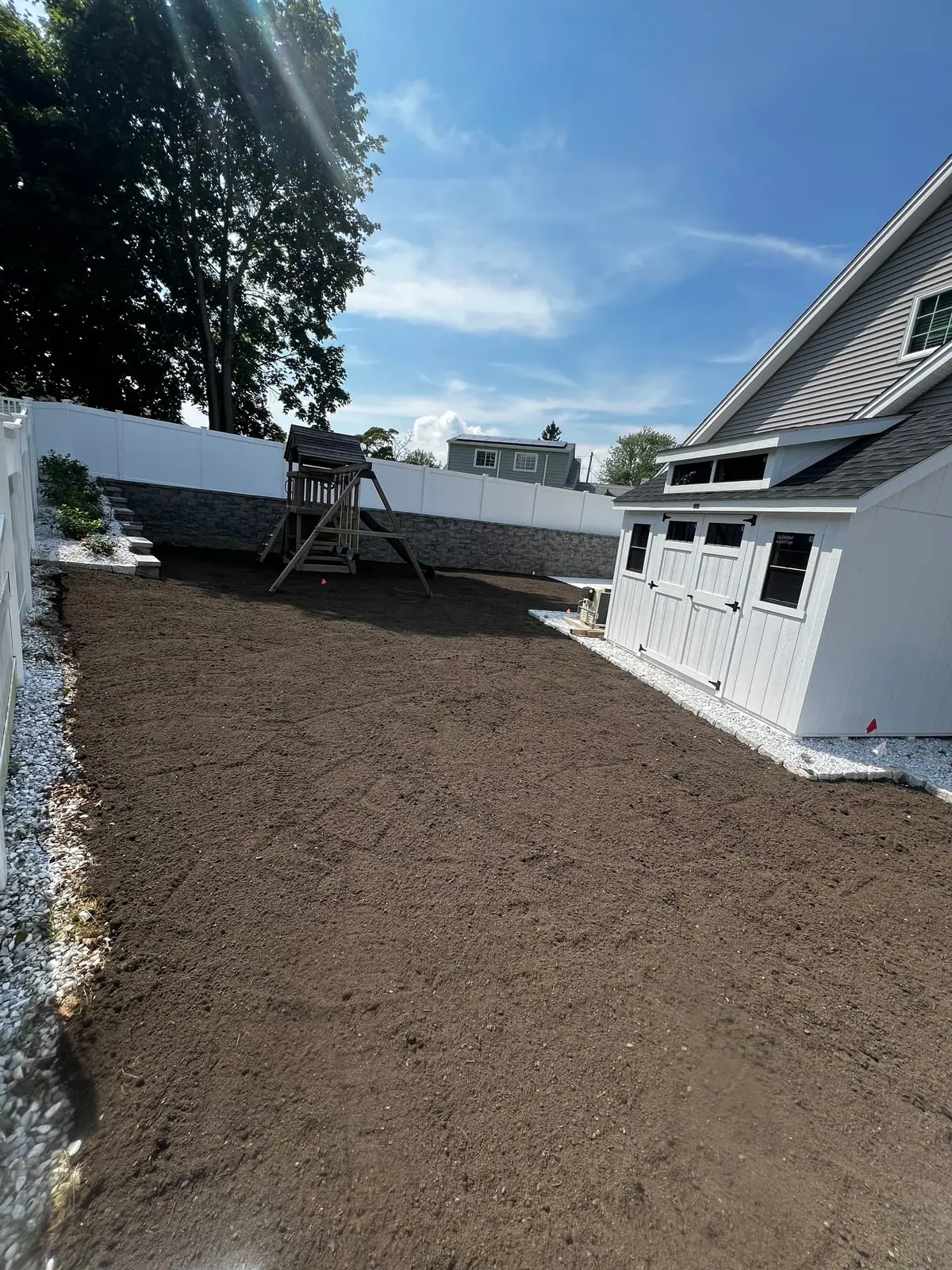 The graded Milford, CT backyard ready for sod — a wide, level bed of raked dirt running past a wooden playset and a stacked-stone wall on one side and a white board-and-batten shed on the other
