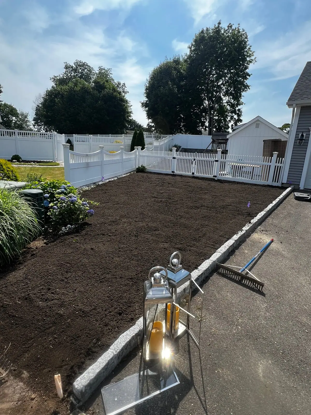 A freshly graded and raked front bed in Milford, CT — dark, level topsoil edged by a granite Belgian-block curb, hydrangeas to one side and a white vinyl fence behind, with a landscape rake resting on the curb