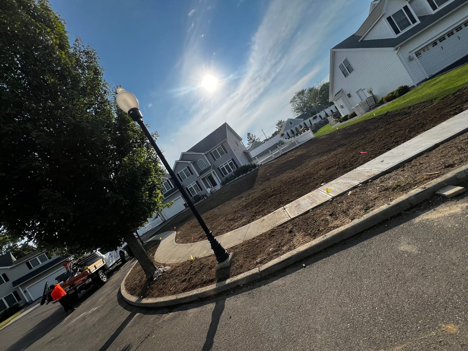 The graded front yard in Milford, CT seen from the cul-de-sac — a sloped bed of dark, level soil above a new concrete sidewalk and granite curb, the gray colonial up the rise, the crew’s dump truck staged at the curb