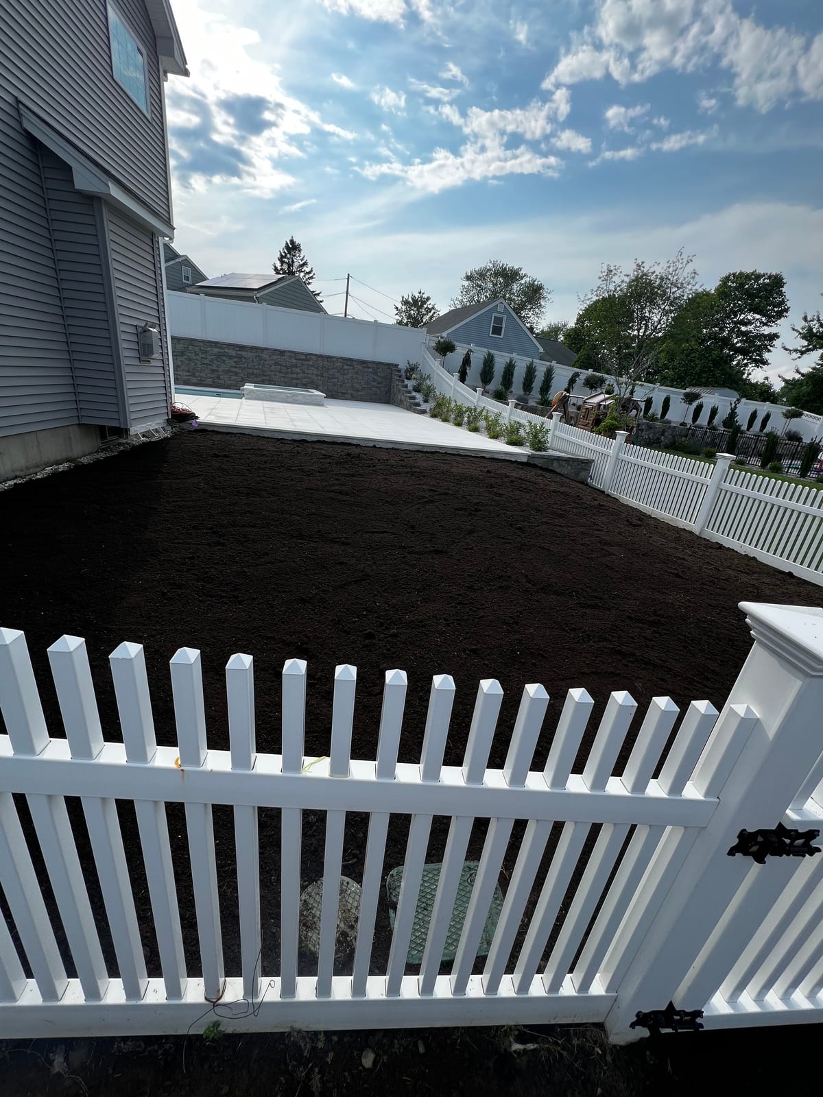 Dark, freshly graded topsoil beside the new pool patio in Milford, CT — a raked bed bordered by a white vinyl picket fence in front and a paver patio and stacked-stone wall behind, ready for sod