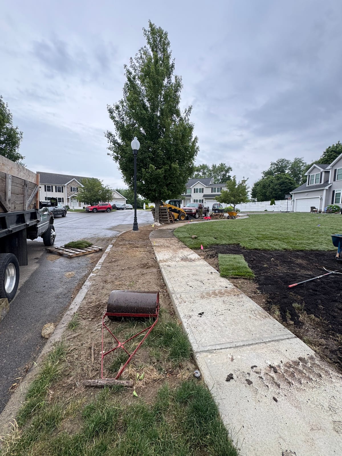 Final front-yard prep in Milford, CT along the sidewalk — a red water-filled lawn roller on the grass strip, dark topsoil raked smooth behind the granite Belgian-block curb, fresh sod started on the right, and pallets of tall fescue staged by the dump truck and skid-steer