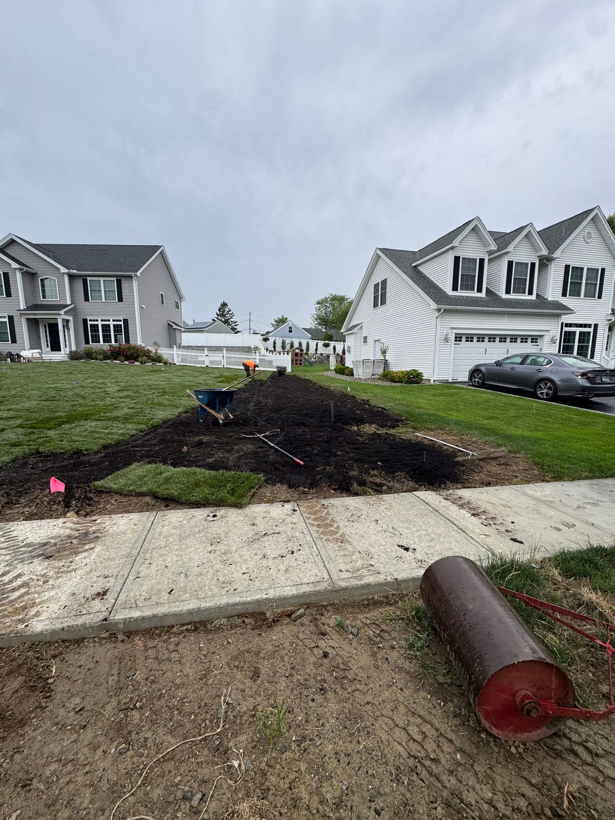 A crew member top-dressing the graded front yard in Milford, CT — spreading and raking dark screened topsoil from a wheelbarrow behind the sidewalk, a water-filled lawn roller on the grass nearby and the first strip of tall fescue sod already down at the left, the gray colonial and a white neighbor's home behind