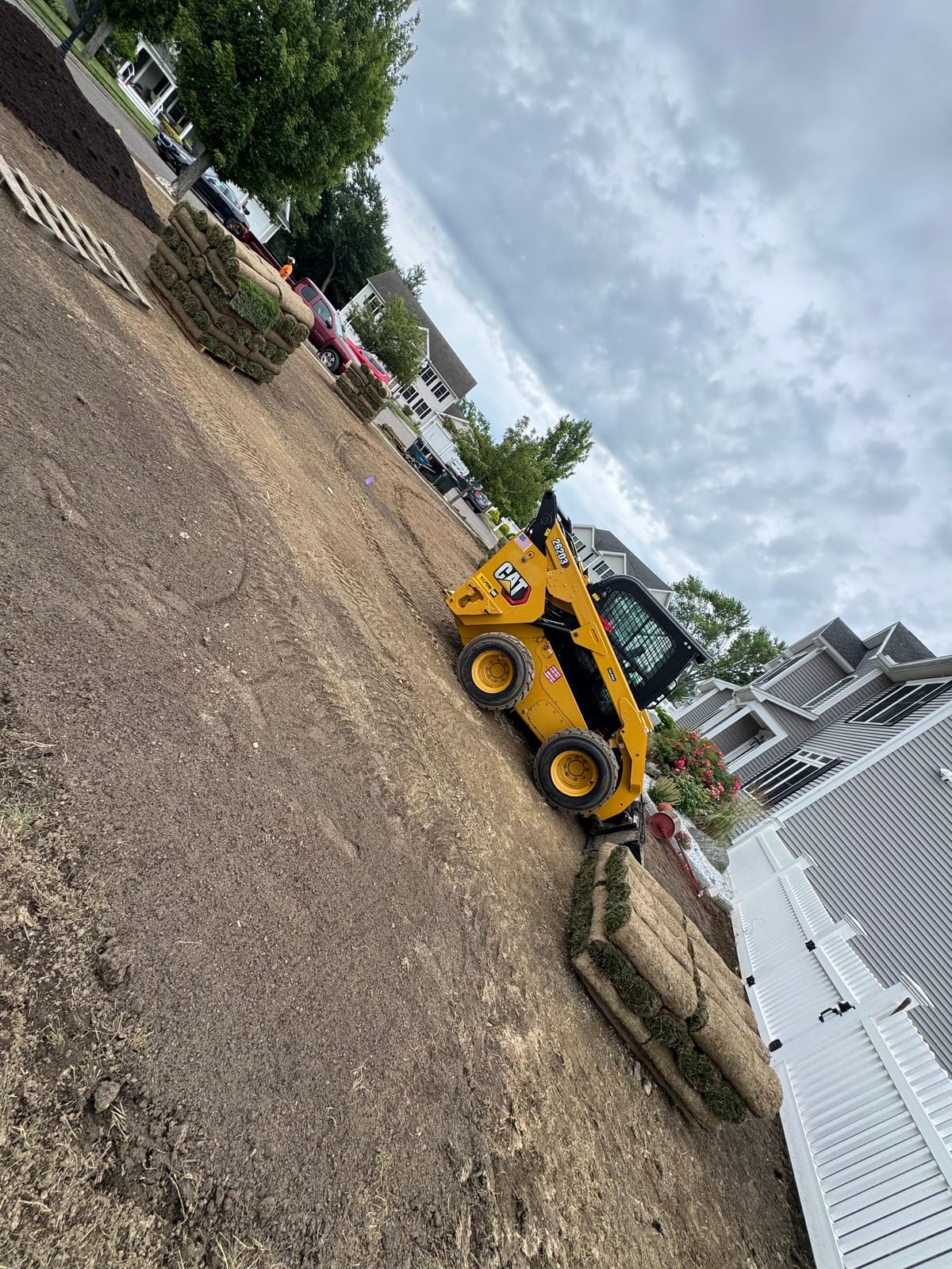 A yellow Cat track loader moving pallets of tall fescue sod across the bare graded front yard in Milford, CT, fresh rolls stacked by the white picket fence and the gray colonial behind