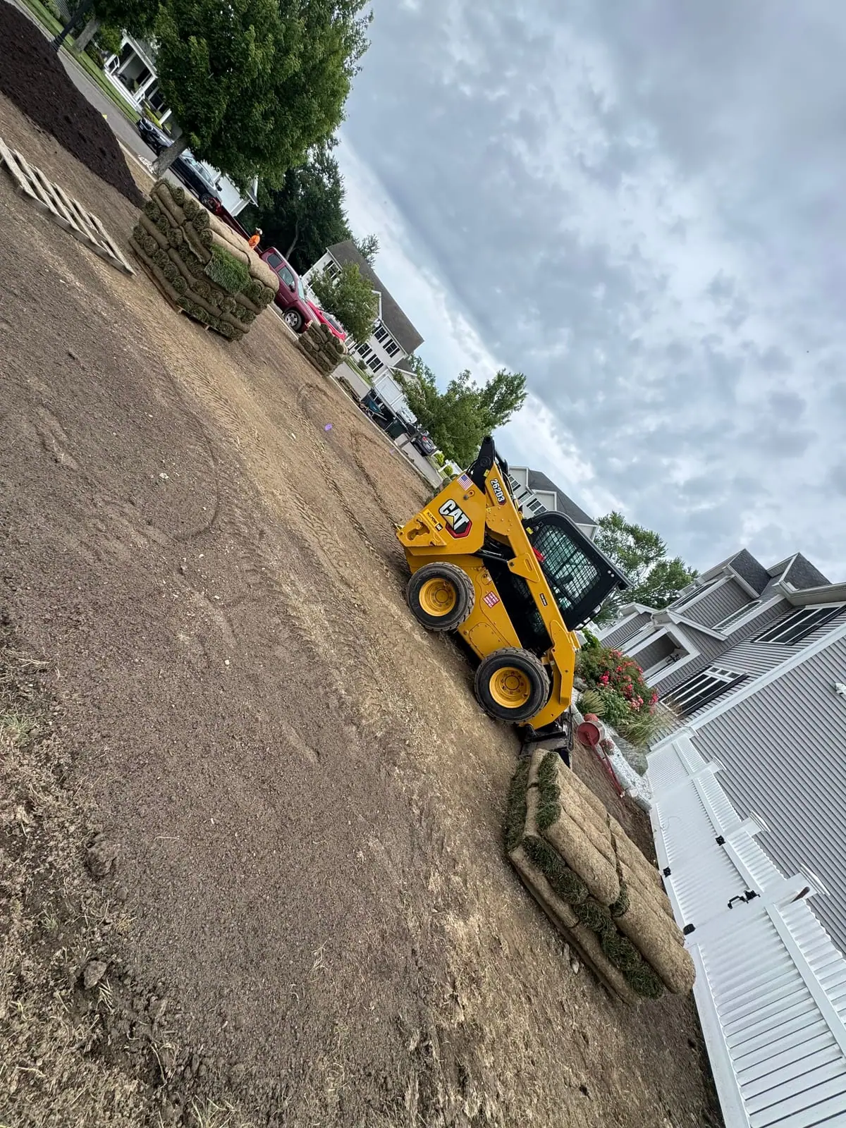 A yellow Cat track loader moving pallets of tall fescue sod across the bare graded front yard in Milford, CT, fresh rolls stacked by the white picket fence and the gray colonial behind