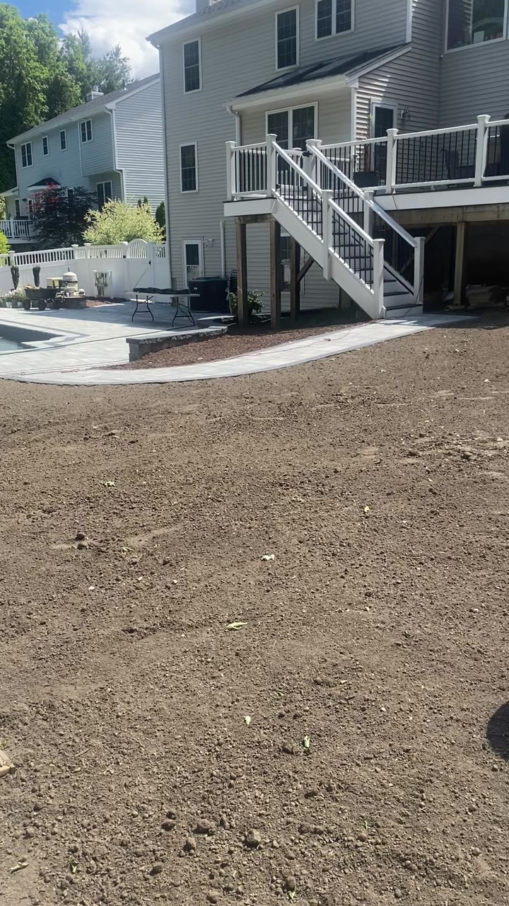 Before a sod installation in Milford, CT — bare graded topsoil across the backyard with a brand-new curved paver patio, inground pool, and raised deck behind it, ready for fresh sod