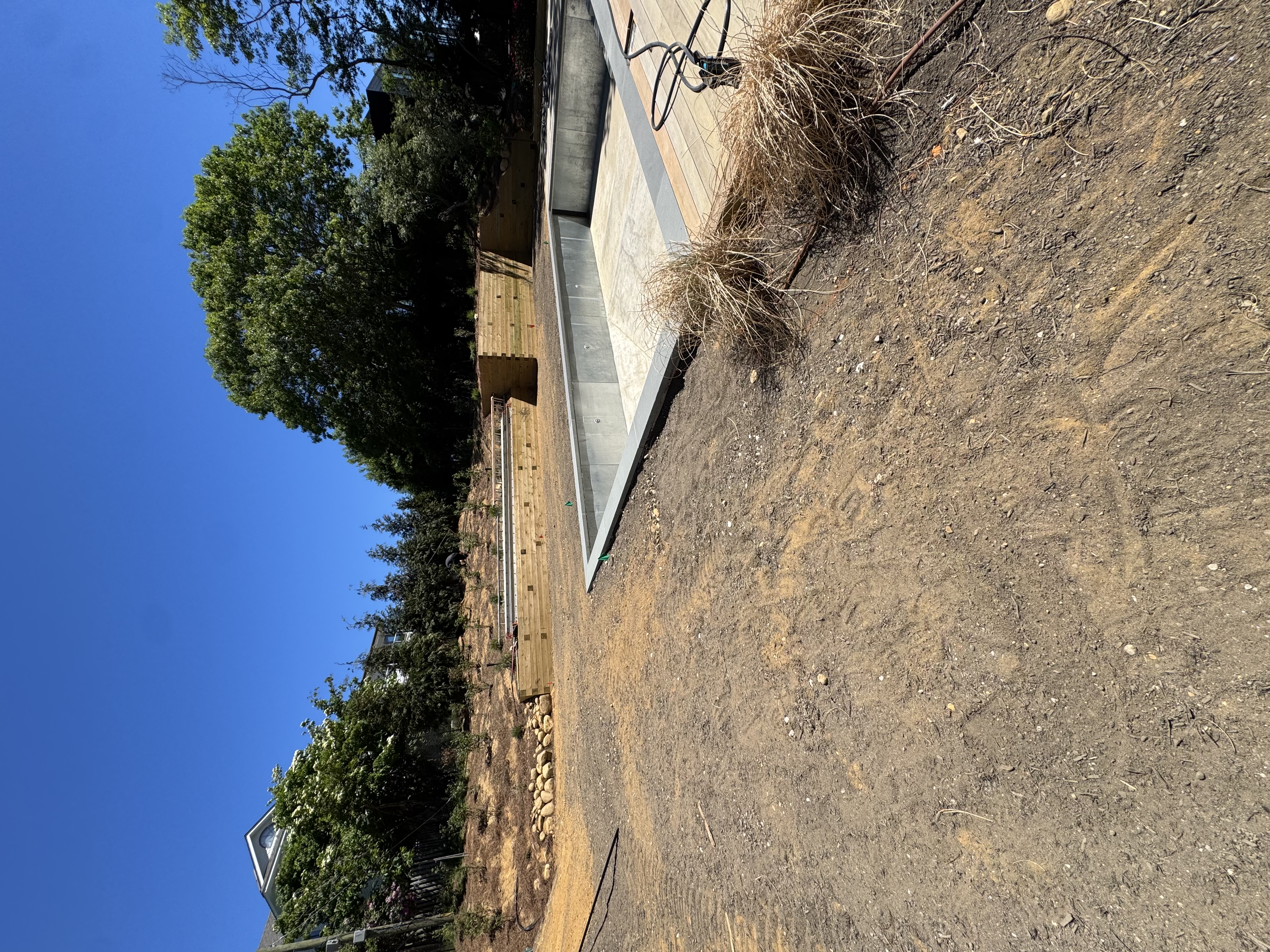 Bare soil and timber retaining-wall terraces wrapping the new pool, graded and ready for fresh sod