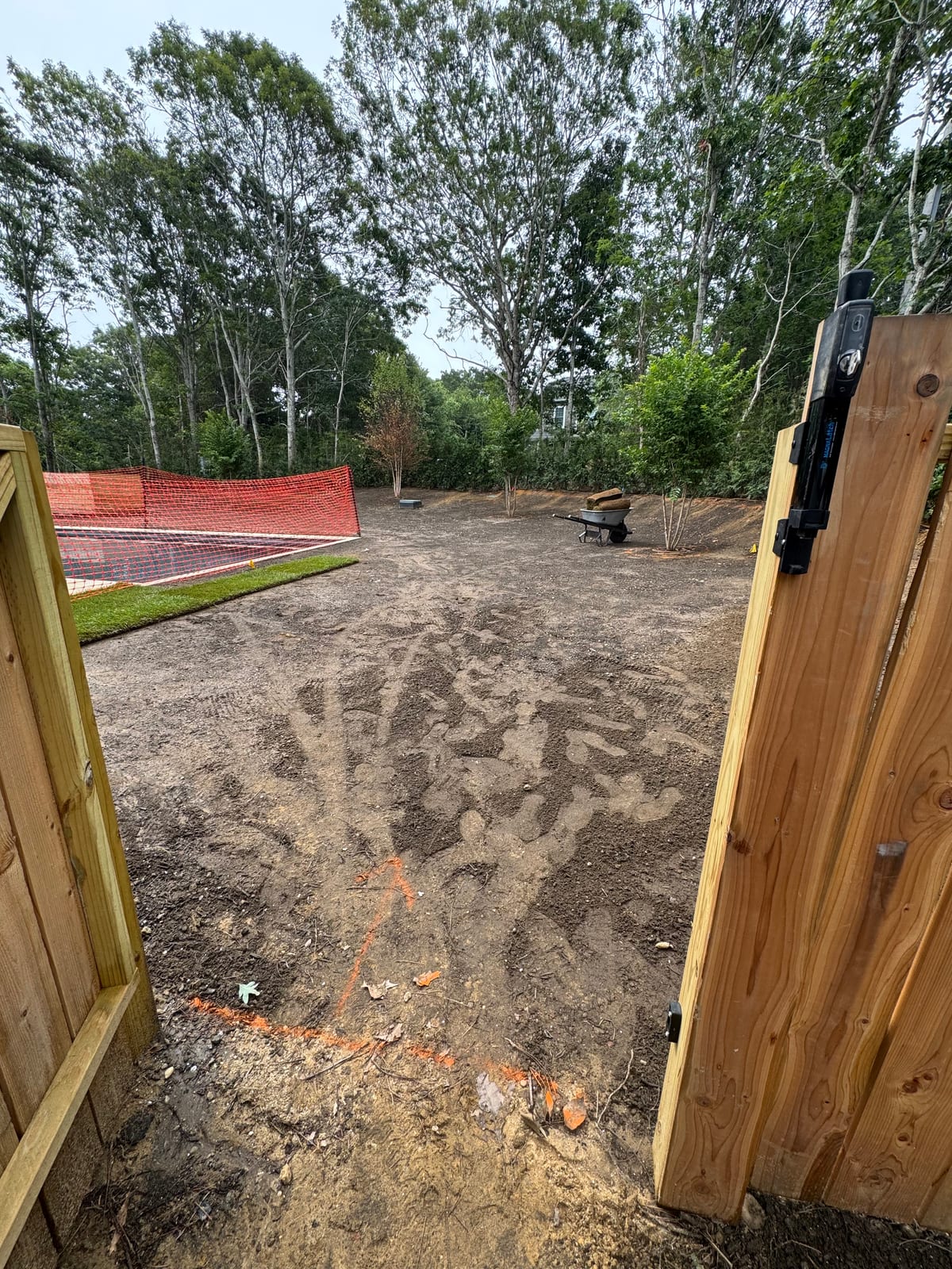 Graded dirt yard seen through the cedar gate before sod installation at the Montauk new build, with the pool and orange safety fencing beyond