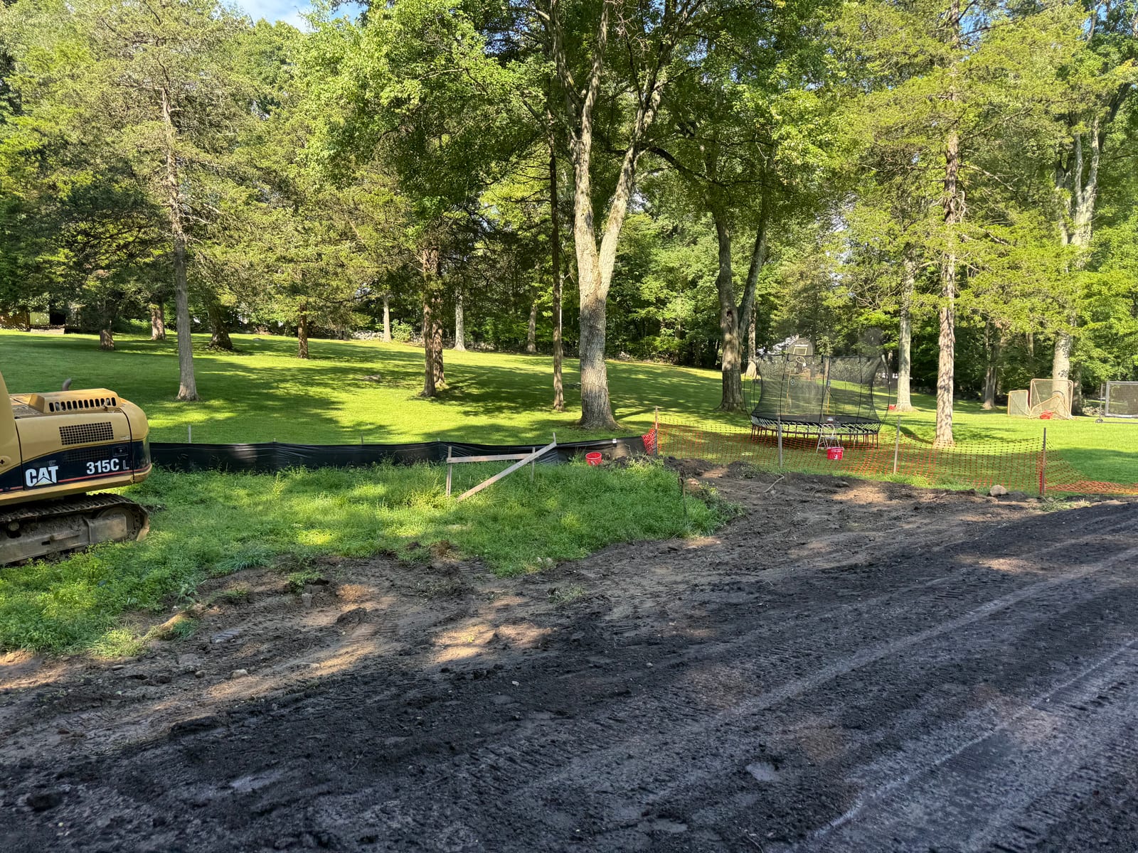 Graded soil meeting the established back lawn at the New Canaan, CT property, with a trampoline and basketball hoop behind orange safety fencing
