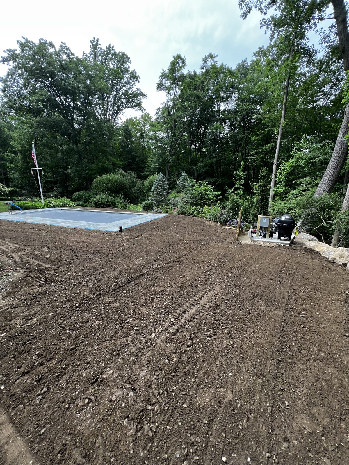 Freshly graded topsoil raked smooth around a new covered pool in New Canaan, CT, with the stone coping taped off and mature plantings framing the yard