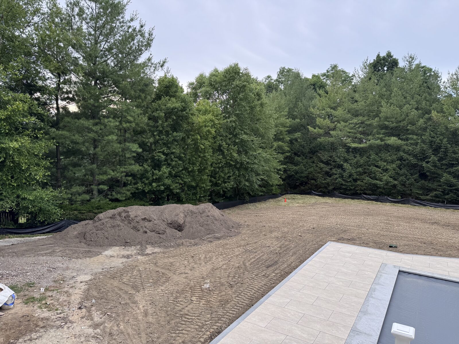 Wide view of a Purchase, NY backyard stripped to dirt after pool construction, with a large topsoil pile and machine tracks across the yard