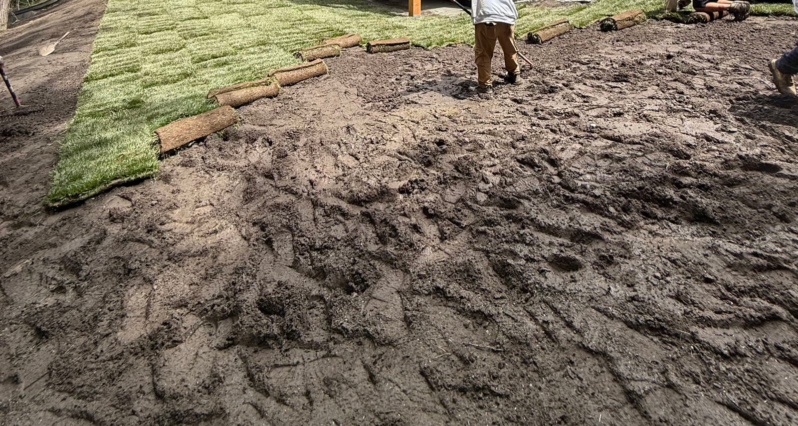 CT Sod crew laying fresh tall fescue sod straight over the mud beside the modern black home in Hartford, VT (Quechee) — green rolls already blanketing one half of the slope while a crew member rakes out the bare, churned ground on the other