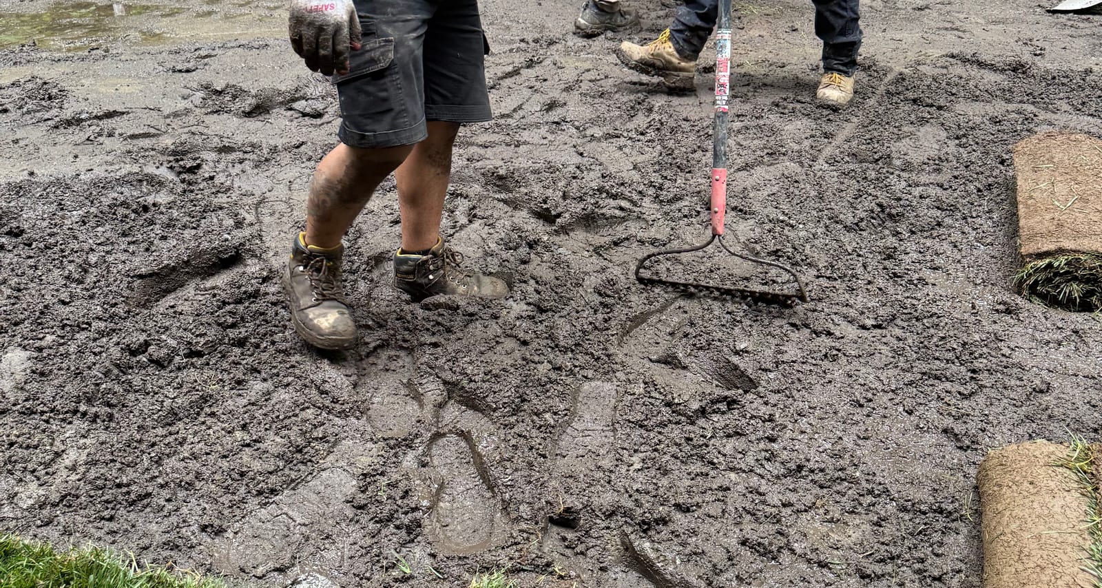 A CT Sod crew member trudging across the deep, churned mud in soaked work boots while the crew rakes the saturated ground smooth by hand — fresh tall fescue rolls and a green sod edge waiting at the side in Hartford, VT (Quechee)