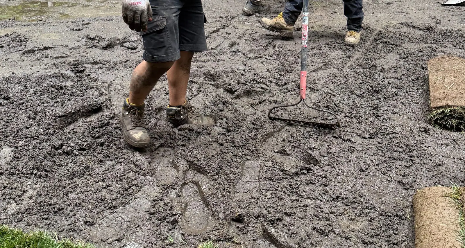 A CT Sod crew member trudging across the deep, churned mud in soaked work boots while the crew rakes the saturated ground smooth by hand, fresh tall fescue rolls and a green sod edge waiting at the side in Hartford, VT (Quechee)