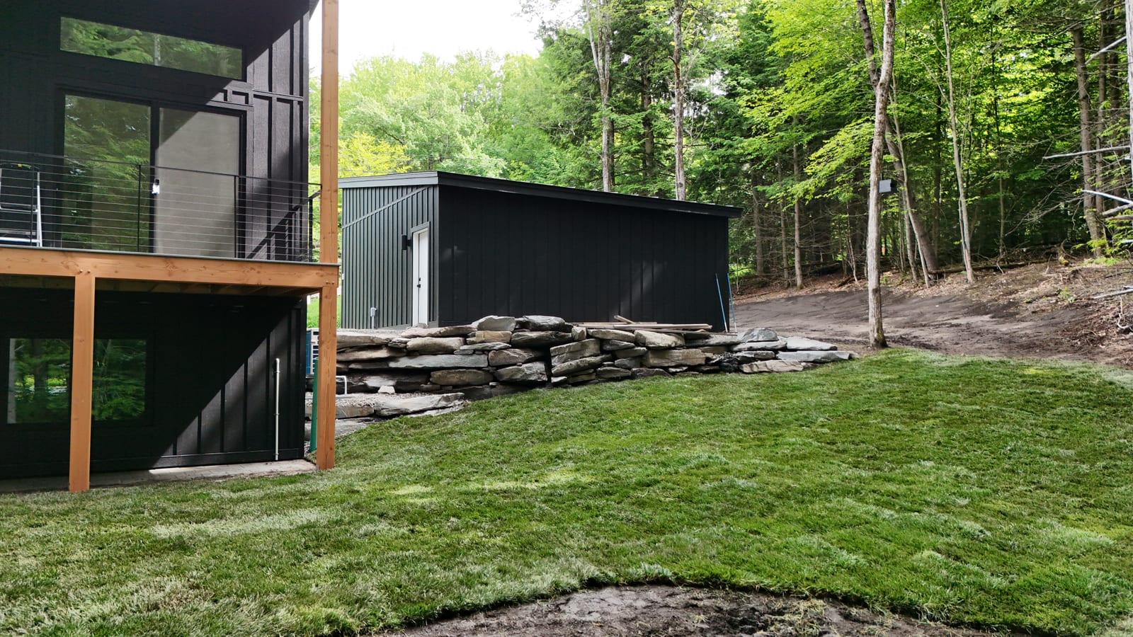 Finished tall fescue lawn beside the home's black outbuilding and a stacked-stone retaining wall, the green slope falling away toward the Vermont woods