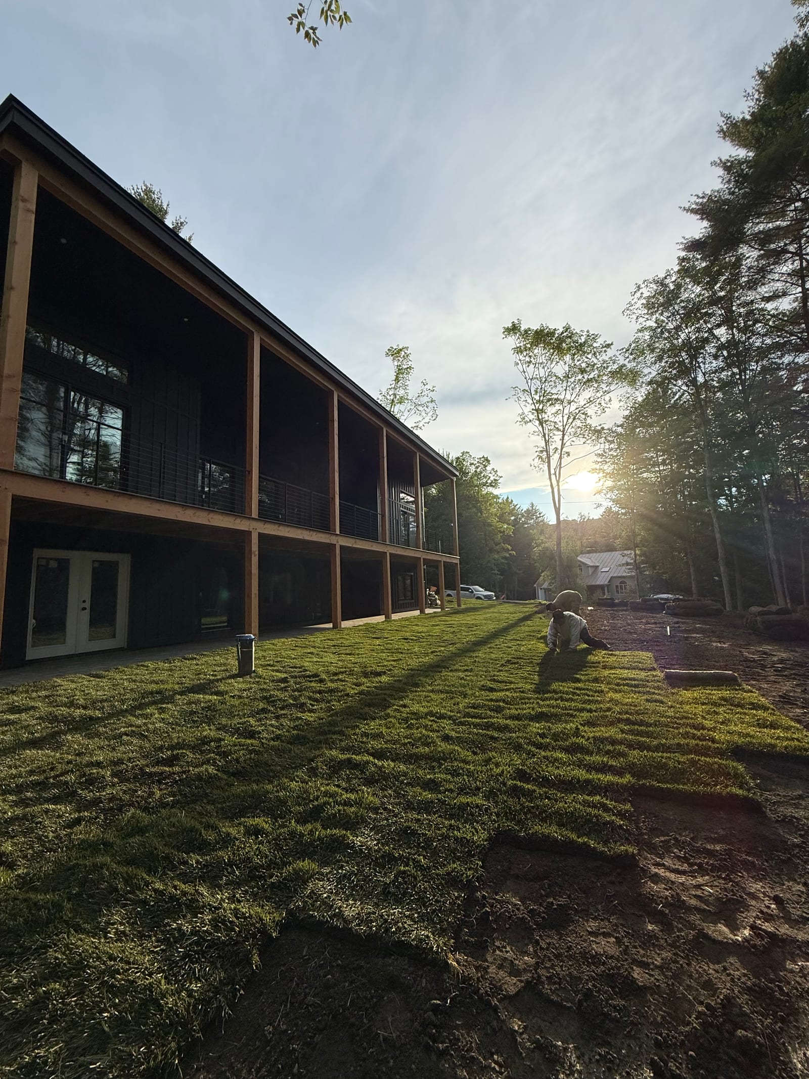 Golden-hour light raking across the finished tall fescue lawn, long shadows stretching down the slope below the home's two-story porch at the end of the longest day