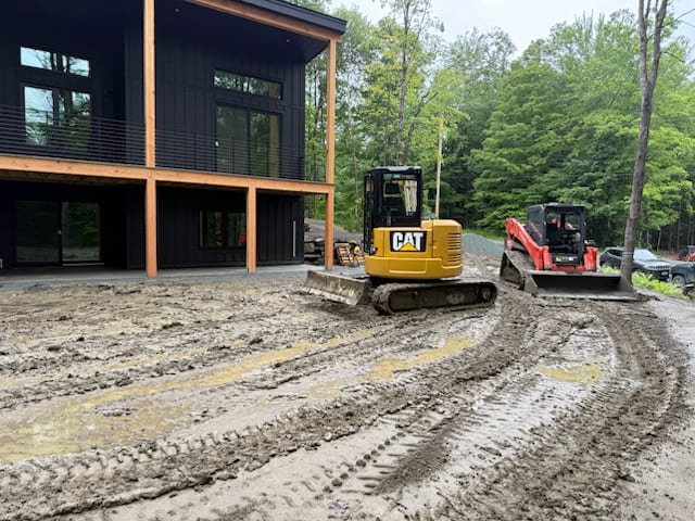 Compact excavator and skid steer regrading the muddy ground in front of the home before sod goes down in Hartford, VT