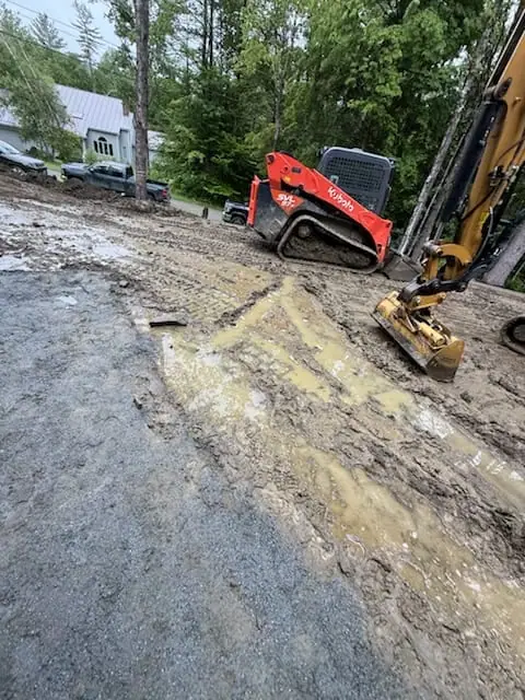 A compact excavator and red skid steer parked on the soupy access drive, the morning's rain still standing in the ruts on the Quechee site