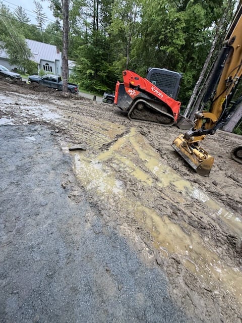 Ground-level view of deep mud and ruts on the Hartford, VT site with a compact skid steer working the saturated ground