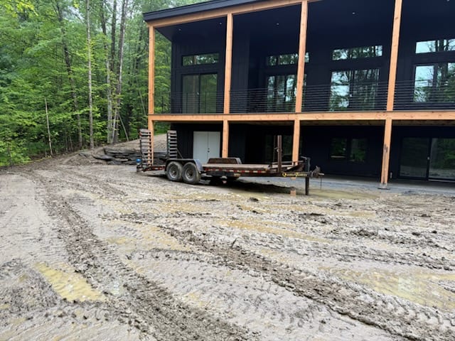 A compact excavator and track loader on the rain-churned, rutted access drive in front of the modern Hartford, VT home, working the saturated ground back to grade