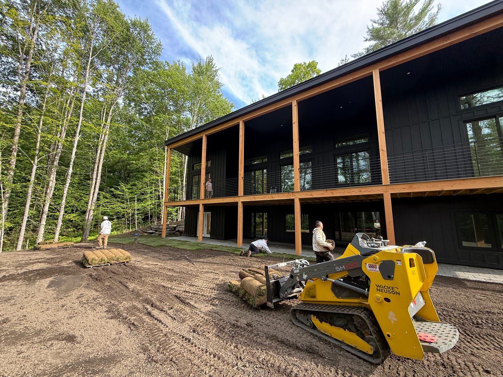 A Wacker Neuson stand-on track loader carrying fresh-cut tall fescue rolls across the yard while the CT Sod crew lays them along the modern black home in Hartford, VT (Quechee), keeping foot traffic off the firmed bed