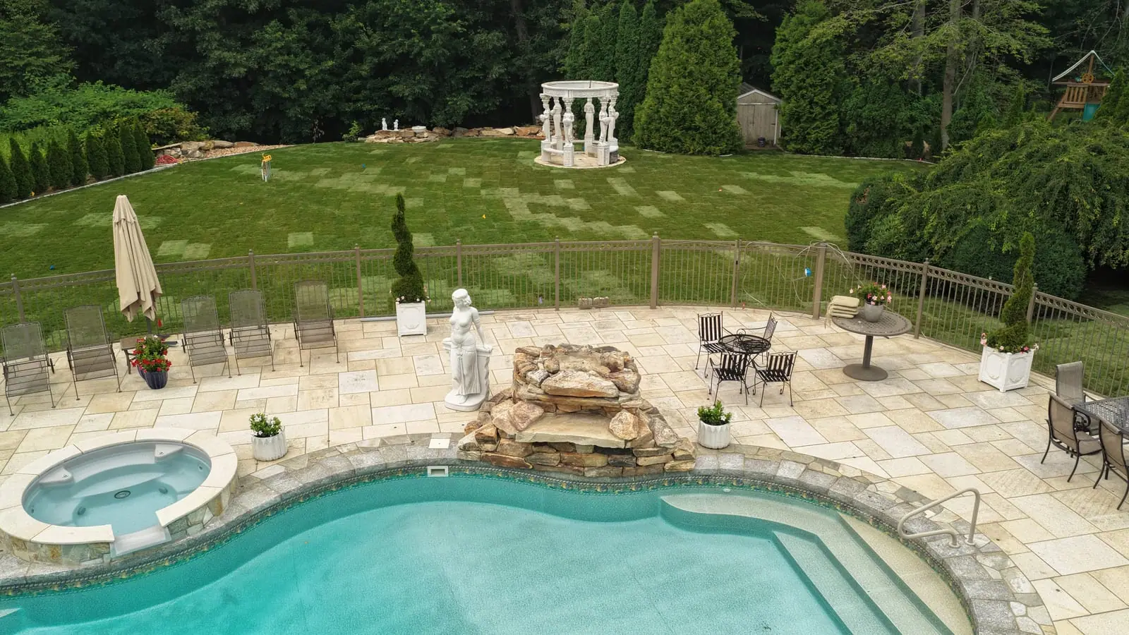 View from the pool patio in Shelton, CT after the sod installation — hot tub, stone waterfall, and statue in the foreground with fresh sod running back to the white gazebo