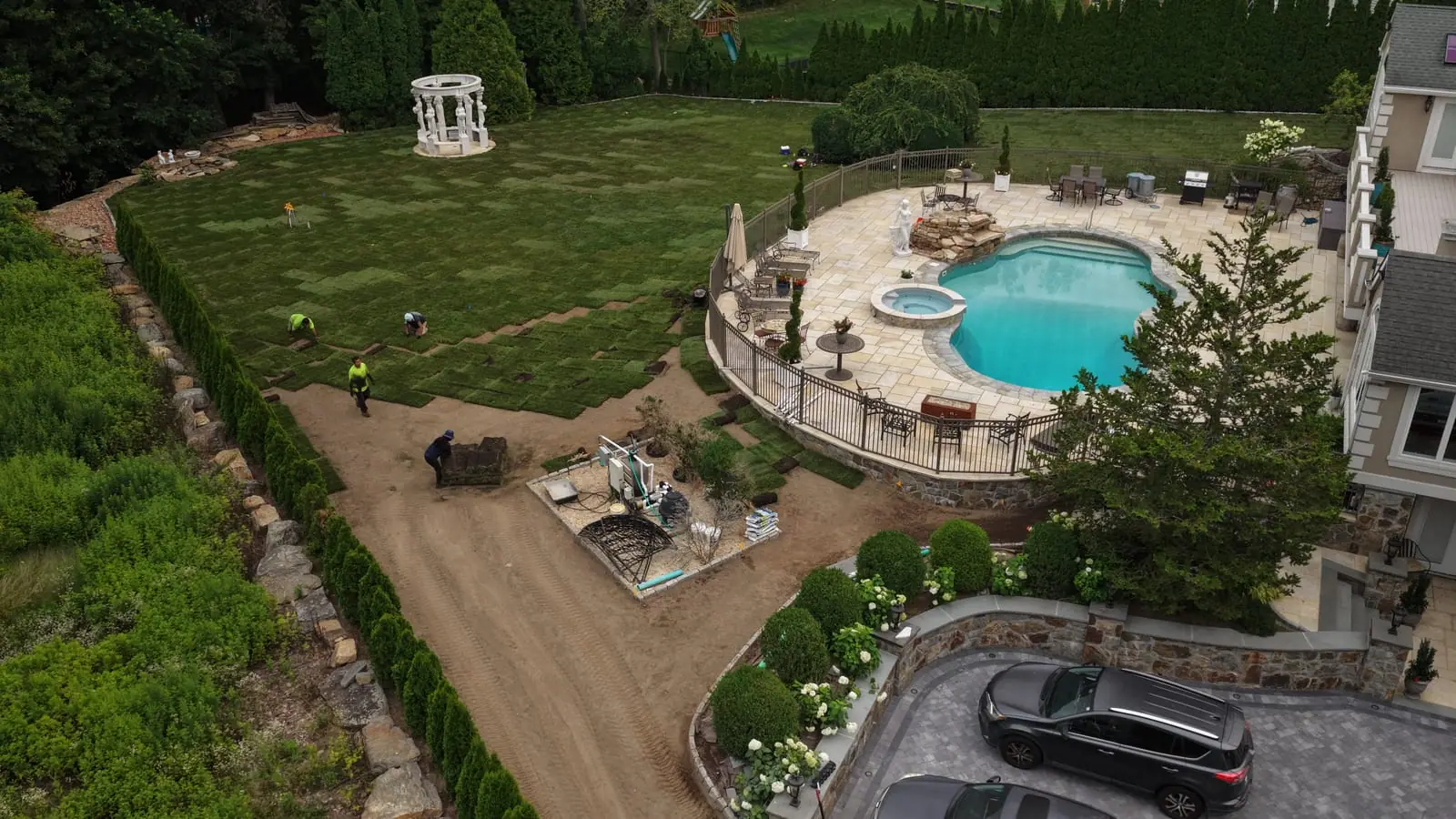 Low aerial of the crew hand-laying sod pieces across the Shelton, CT back yard slope — staged stacks just ahead of the working edge, with the pool patio and statue beyond