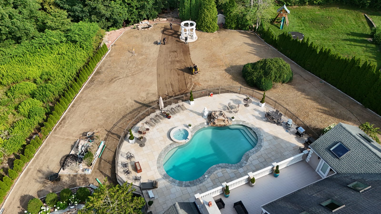 Aerial view over the new pool and spa in Shelton, CT, looking across 17,000 square feet of graded bare dirt with a white gazebo, a compact track loader, and the crew spreading topsoil