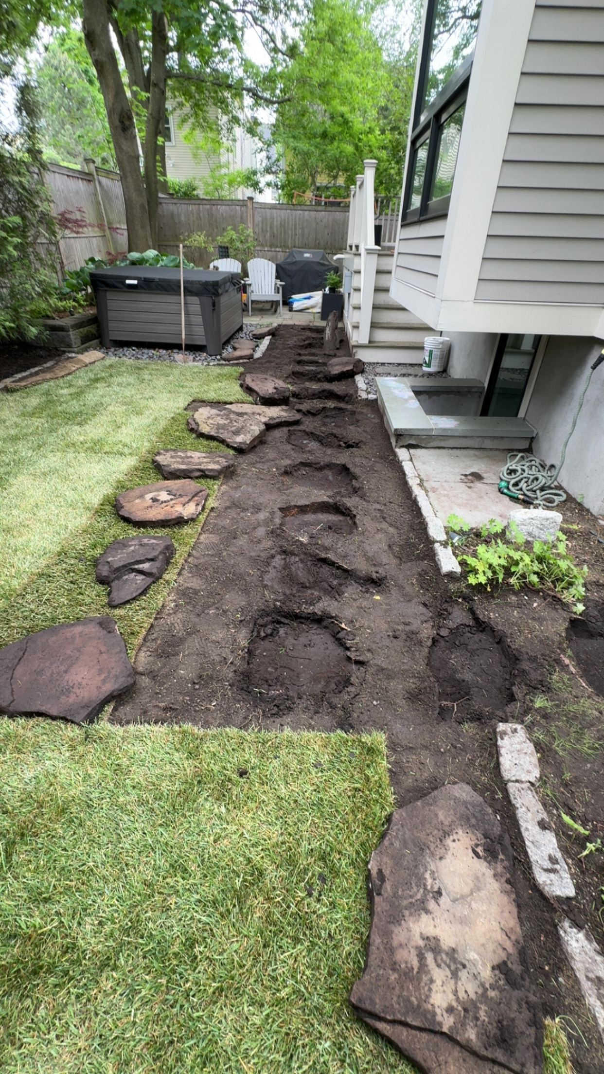 Cambridge backyard mid-prep — stepping stones lifted out, dark soil exposed along the foundation, ready for grading and sod