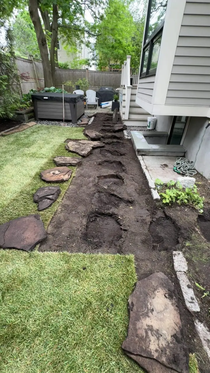 Cambridge, MA backyard mid-prep — stepping stones lifted out and dark soil exposed along the foundation, ready for grading and sod