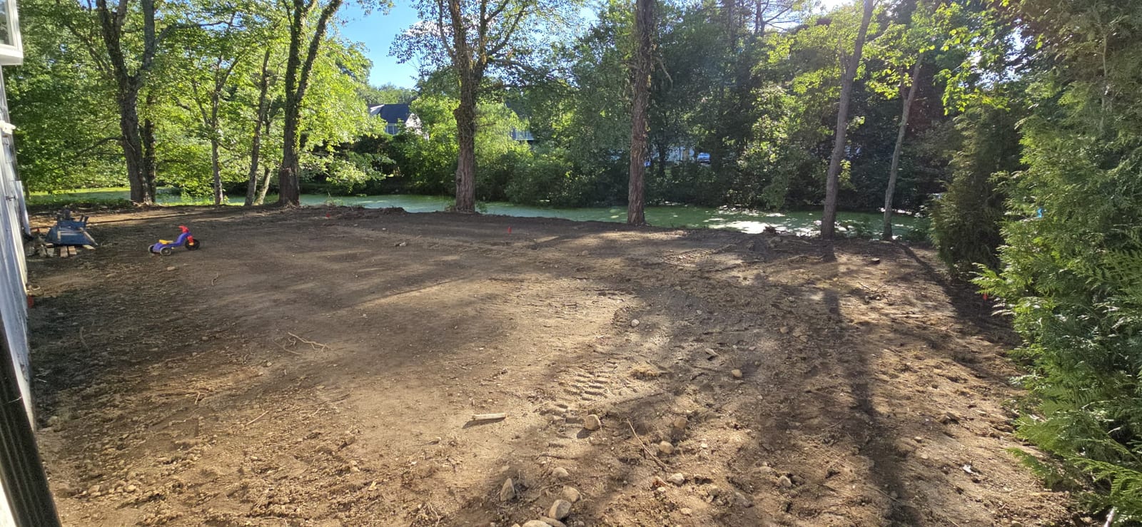 Graded bare-dirt backyard in Stamford, CT sloping down toward a tree-lined pond, raked smooth and ready for sod