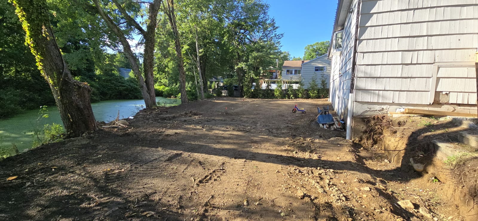 Graded side yard beside a white house in Stamford, CT, bare soil raked level and running down toward the water, ready for Kentucky bluegrass sod