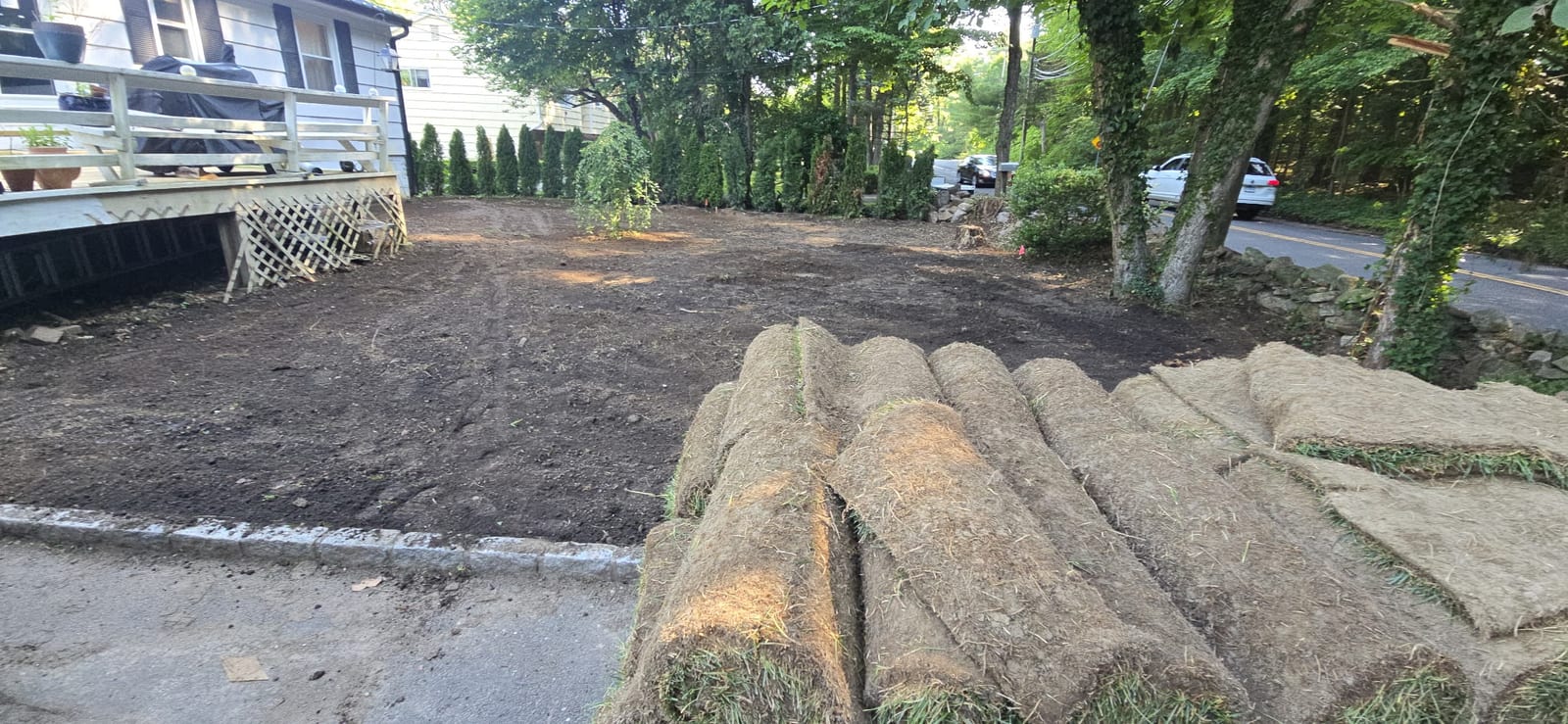 Stacks of fresh-cut Kentucky bluegrass sod rolls staged in the driveway of a Stamford, CT home, with the graded backyard and deck behind
