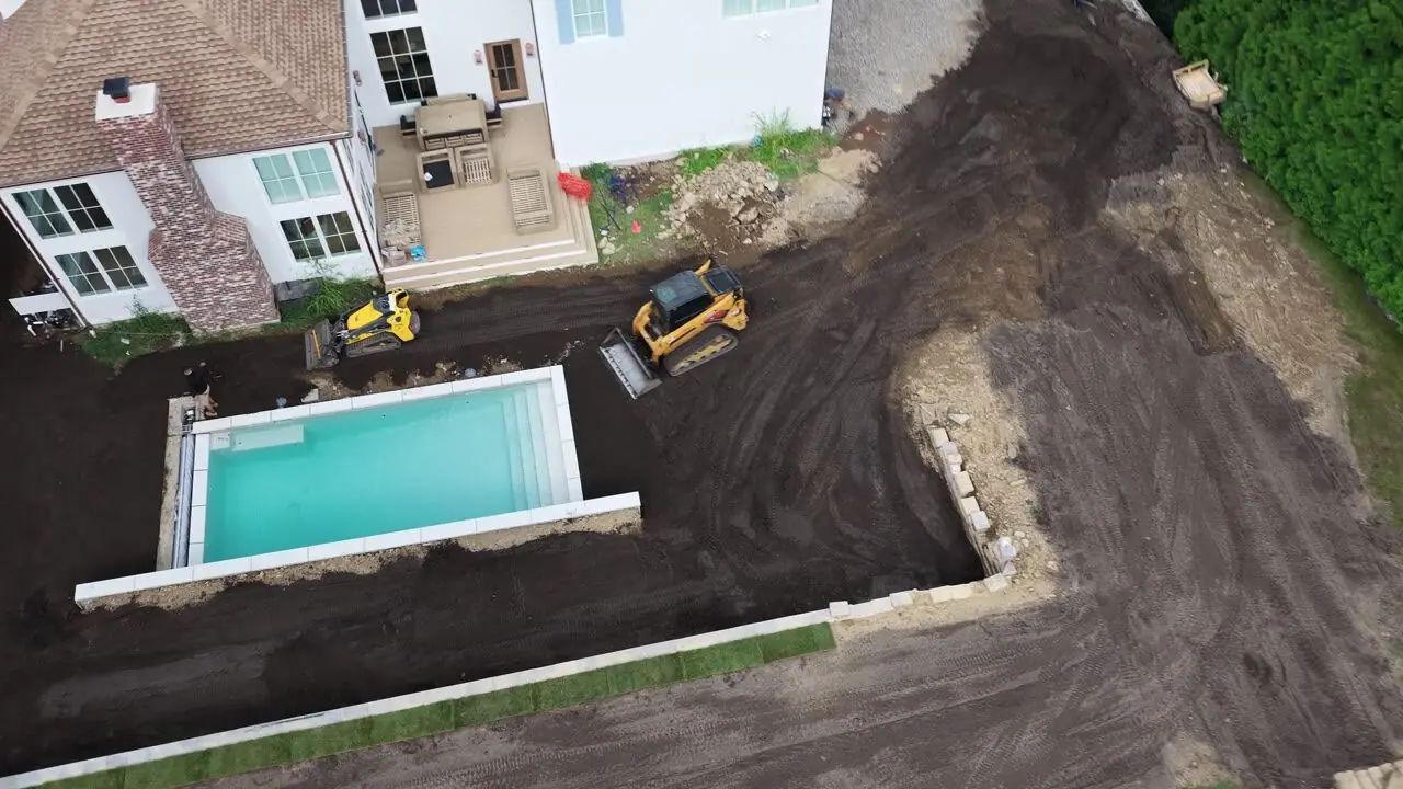 3:55 p.m. over the finished grade at the pool wall — pallets staged on the driveway while the loader works the side yard