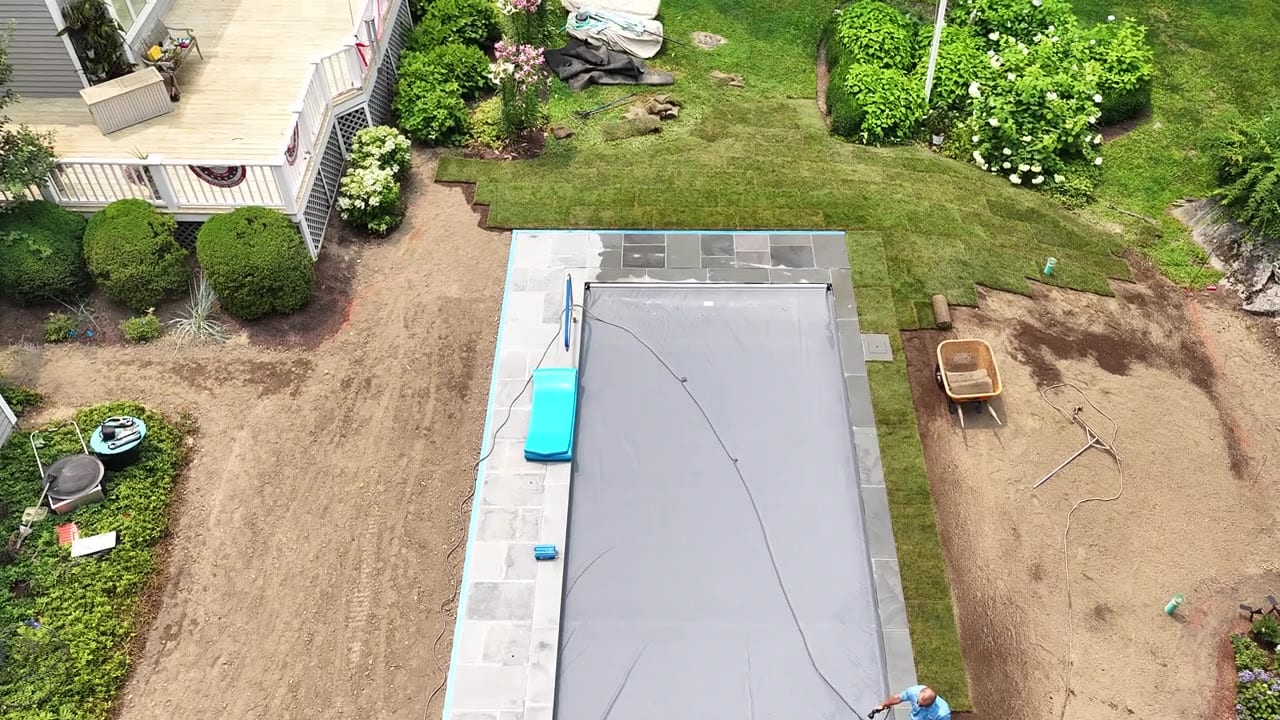 Install day from above — the first rolls of fresh-cut sod going down across the graded yard, working toward the new pool and coping