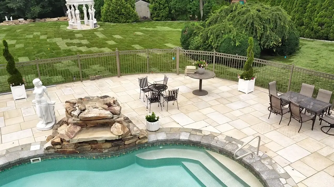 Low drone pass across the Shelton, CT pool patio — dining sets, spa, and stone waterfall with the new lawn beyond the fence