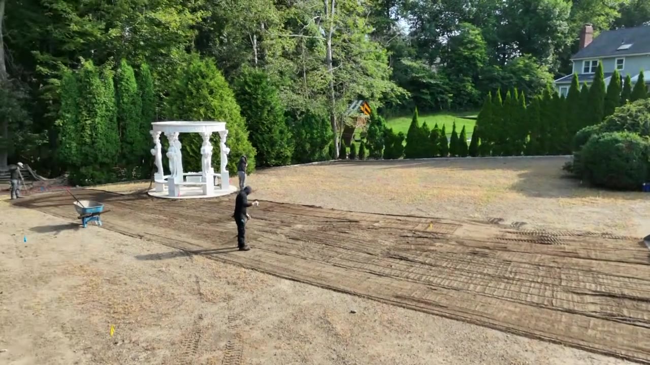 From the pool fence up over the yard — the crew rakes the fresh topsoil smooth around the gazebo while the track loader keeps material coming