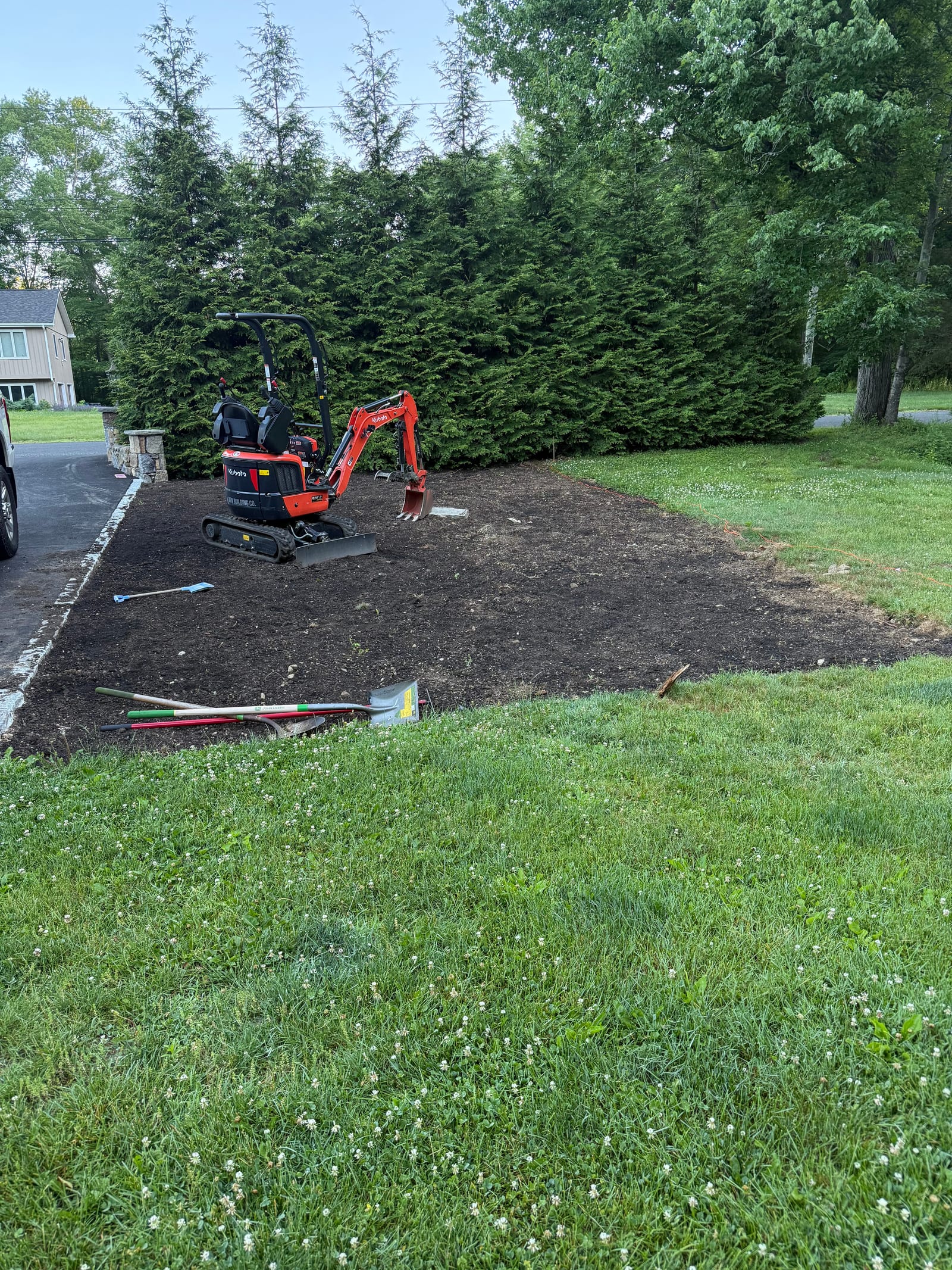 Finish prep for a sod installation in Easton, CT — a compact excavator on freshly graded bare soil abutting the driveway, hand tools staged, with a tall arborvitae hedge behind