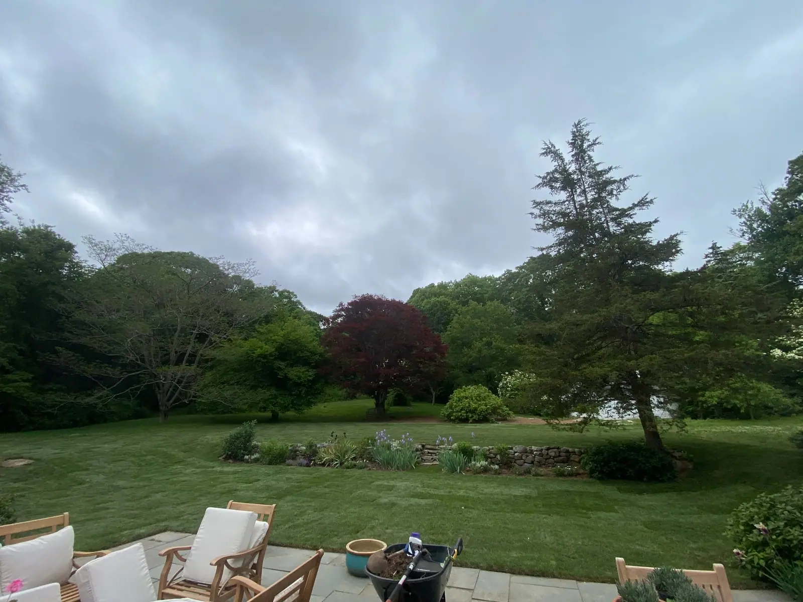 A finished green sod lawn seen from a stone patio with teak chairs and a wheelbarrow, leading out to perennial beds, a stone retaining wall, and trees